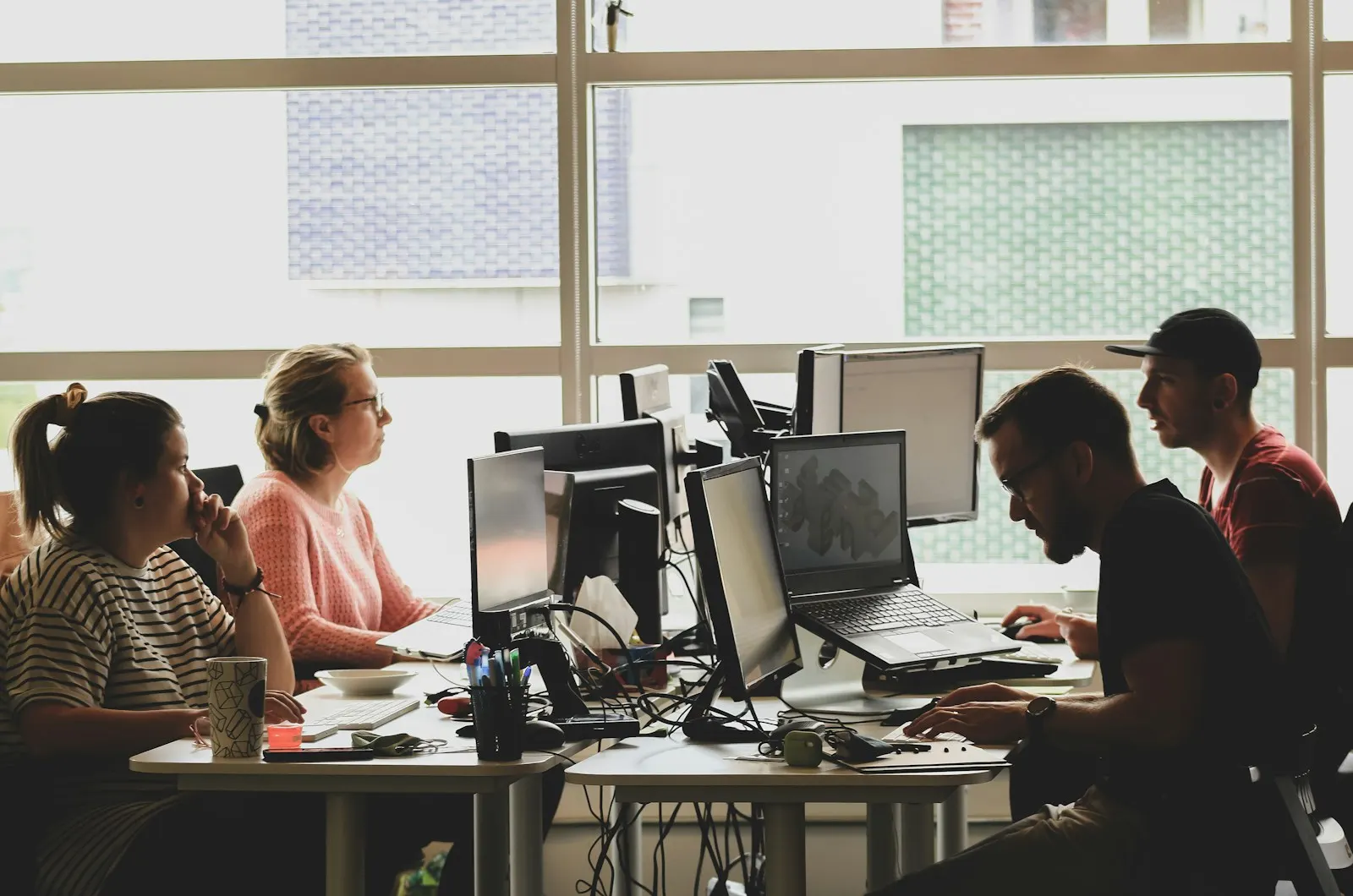 Four people working at computers in a sunlit office workspace — illustrative image